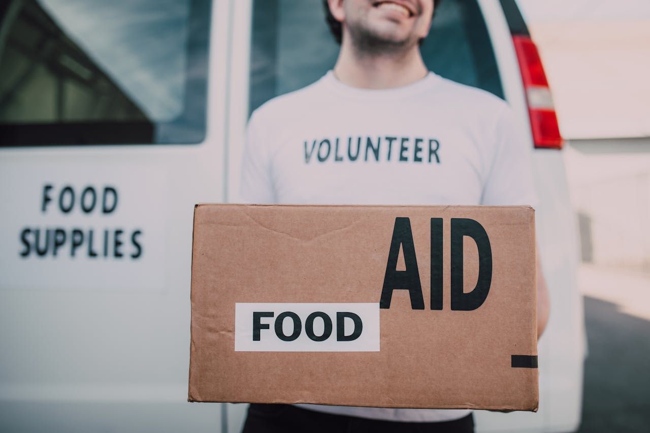 The Art of Drawing Readers In: Your attractive post title goes here Smiling volunteer holding a box labeled 'Food Aid' outside a van with 'Food Supplies' sign.